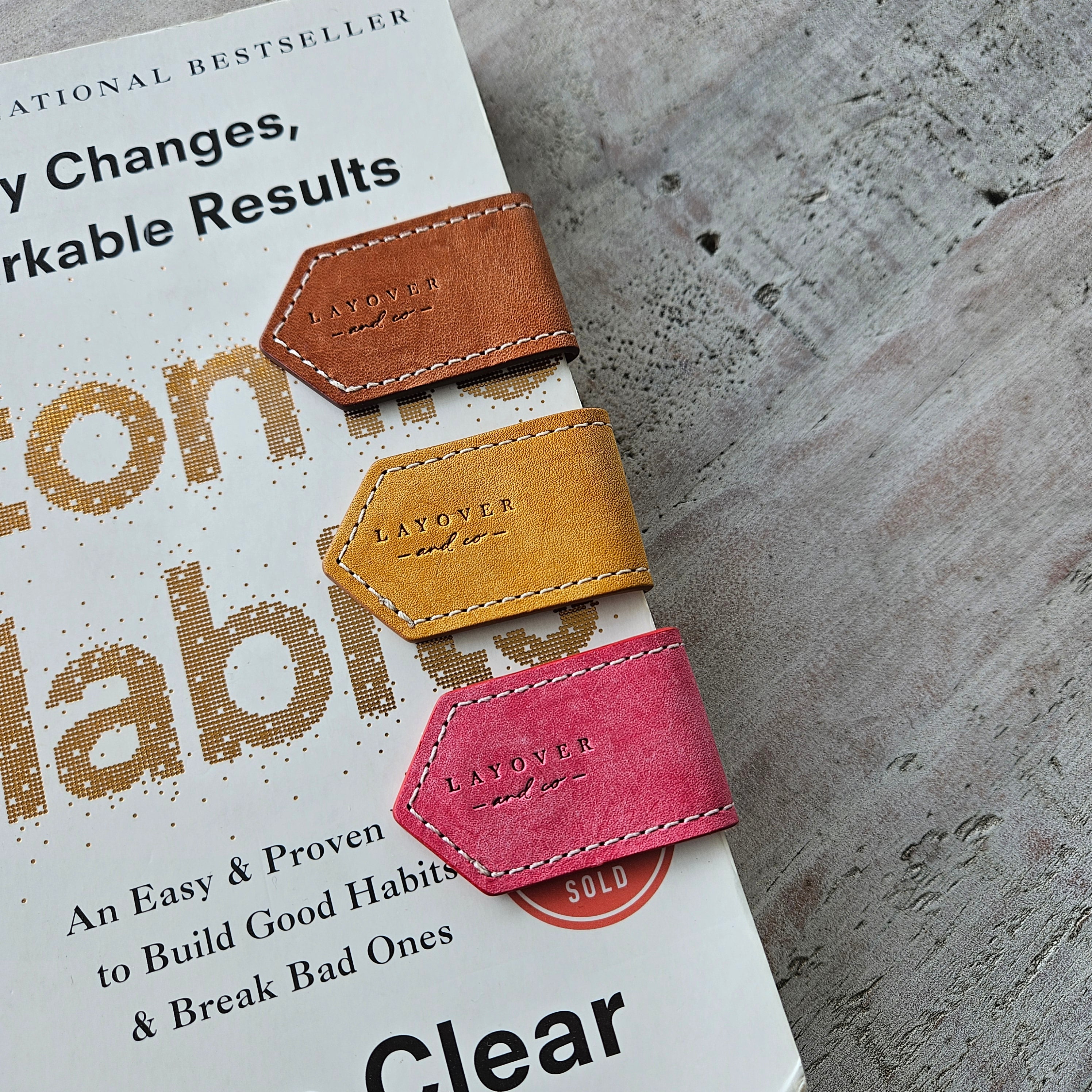 Front-angle view of brown, yellow, and pink leather bookmarks placed inside an open book, showcasing the logo on the bookmarks along with their high-quality, handmade design and functionality in a real-world setting.