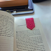 Pink leather bookmark placed inside an open book, surrounded by a computer monitor and desk accessories, highlighting its functionality and elegant design in a real-world setting.
