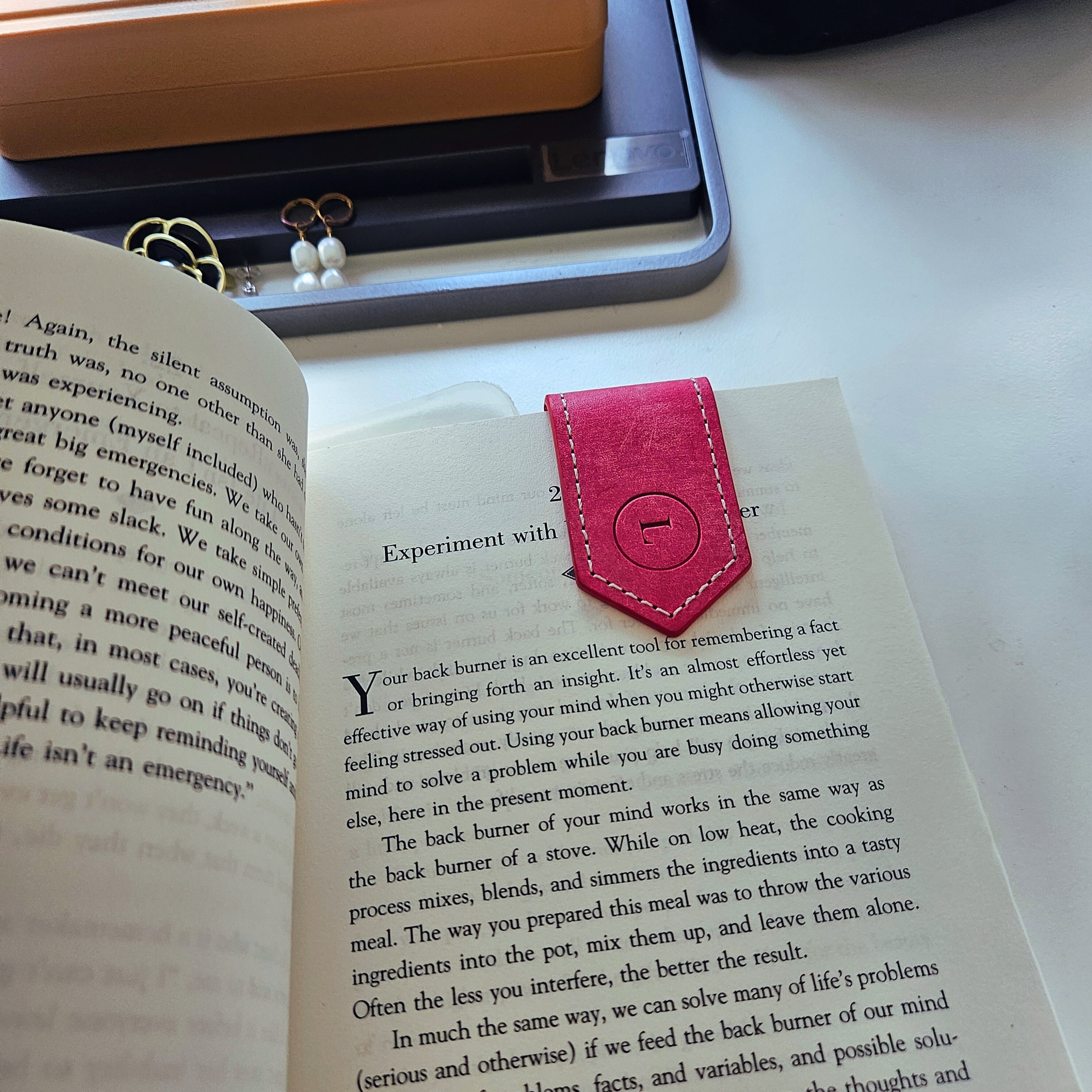 Pink leather bookmark placed inside an open book, surrounded by a computer monitor and desk accessories, highlighting its functionality and elegant design in a real-world setting.