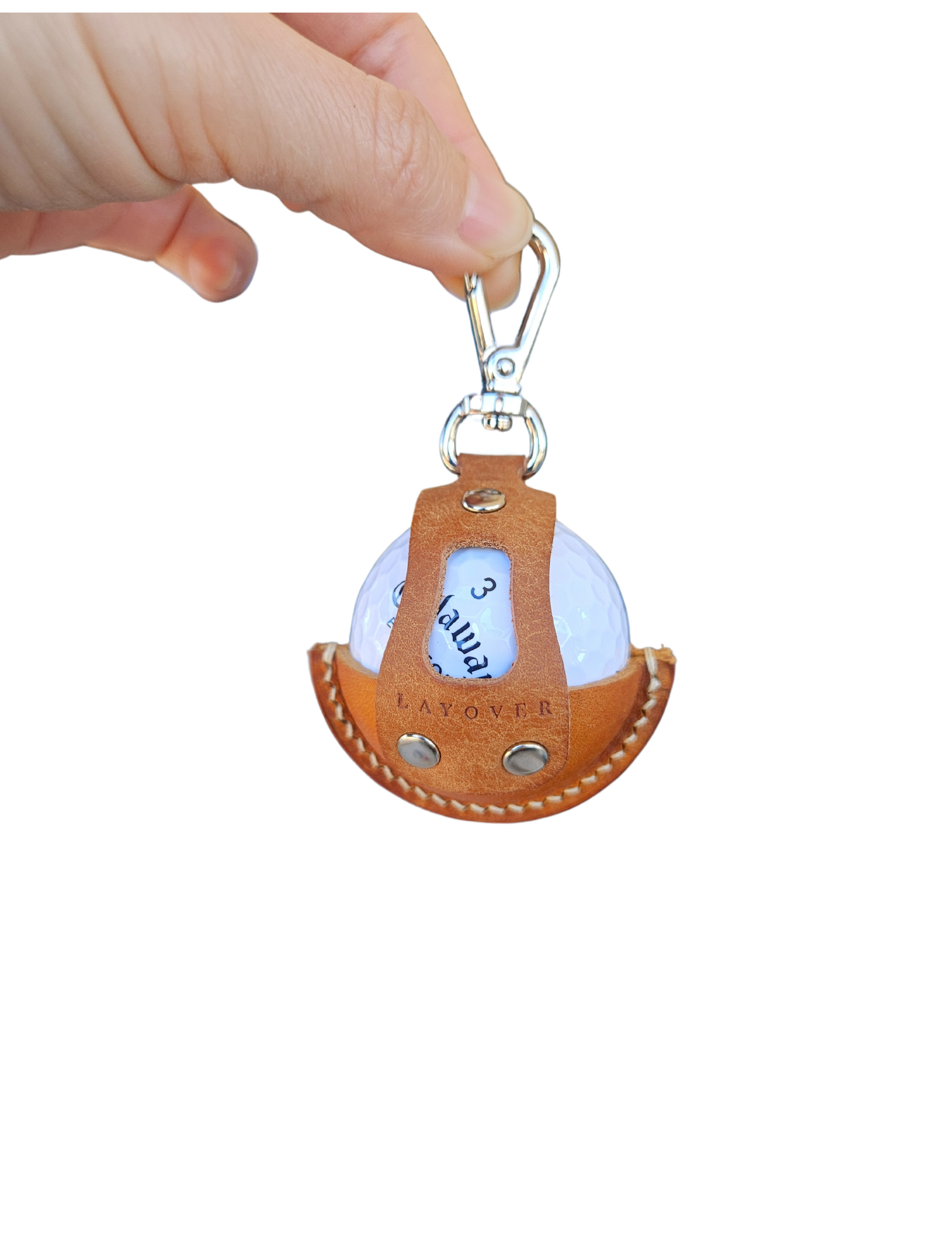 A close-up of a brown leather golf ball holder with visible stitching and metallic rivets, held by a hand. The holder features a secure clip at the top and an open-front design showcasing a white golf ball inside.