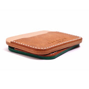 A slim, handcrafted leather card holder featuring a natural tan and brown exterior with white stitching and a green leather layer visible at the bottom. The minimalist design showcases Layover and Co.'s signature craftsmanship.